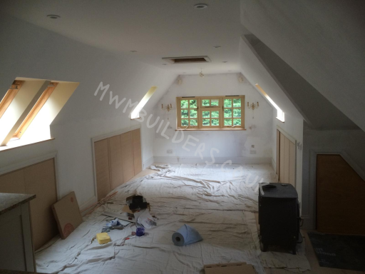 Empty attic room with sloped ceilings, wooden windows, and scattered paint supplies.